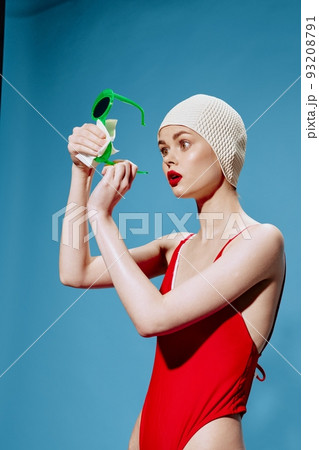 An enthusiastic shocked unusual girl in a swimming cap and a red swimsuit wipes glasses in the studio on a blue background 93208791