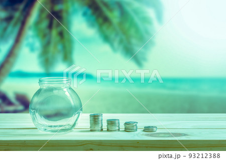 Financial concept, Empty glass bottle to show the money spent on vacation or holidays and stack coins to show decrease of money on wooden table, coconut tree and beach blurred background. 93212388