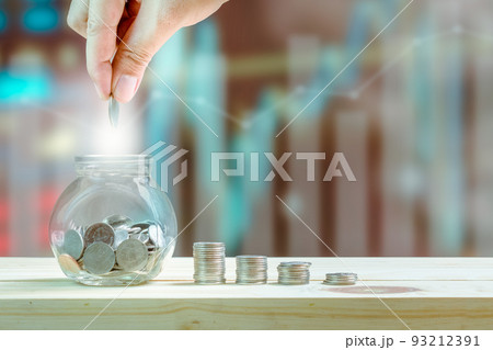 Saving money and investment concept, hand putting coin in glass bottle for savings and stack coins to show increase of savings money on wooden table, graphs and index blurred background. 93212391