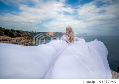 Blonde with long hair on a sunny seashore in a white flowing dress, rear view, silk fabric waving in the wind. Against the backdrop of the blue sky and mountains on the seashore. Blonde with long hair on a sunny seashore in a white flowing dress, rear view, silk fabric waving in the wind. Against the backdrop of the blue sky and mountains on the seashore. 93217943
