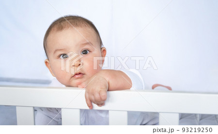 Cute portrait of a baby in a crib close-up. A child in white clothes on white underwear. Tenderness and care, children's problems. 93219250