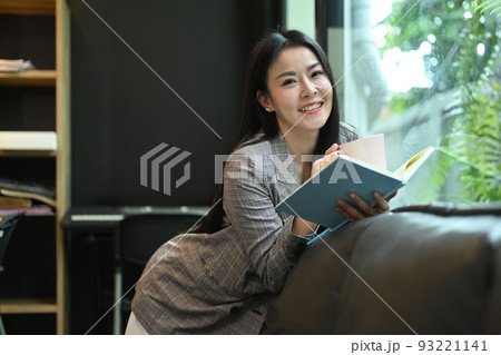 Cheerful asian working woman relaxing on sofa in her bright modern office and reading book Cheerful asian working woman relaxing on sofa in her bright modern office and reading book 93221141