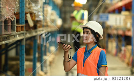 Female warehouse worker wearing safety hard hat and vest scanning barcode on box in a large warehouse Female warehouse worker wearing safety hard hat and vest scanning barcode on box in a large warehouse 93221203