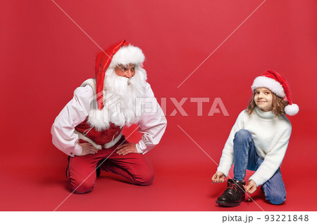 Cute little girl in Christmas hat trying on a boot with Santa against red studio background. Isolate Cute little girl in Christmas hat trying on a boot with Santa against red studio background. Isolate 93221848