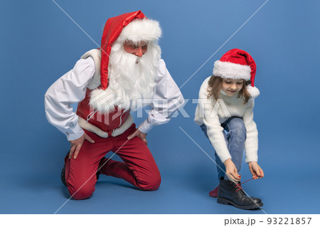 Cute little girl in Christmas hat trying on a boot with Santa against blue studio background. Isolate 93221857