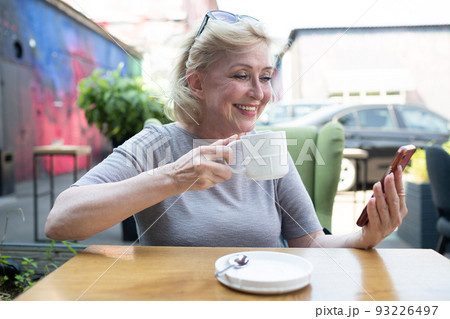 Elderly woman drink coffee or tea drink at the outdoors cafe and using mobile phone Elderly woman drink coffee or tea drink at the outdoors cafe and using mobile phone 93226497