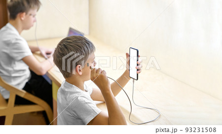 Schoolboys use electronic devices connected directly to head. Younger brother connects cable to smartphone while elder sibling plays game via laptop 93231508
