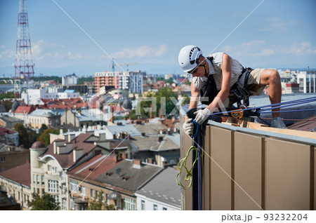Industrial mountaineering worker in safety helmet crouching down on building roof and adjusting climbing rope. Man using safety lifting equipment outdoors with sky and buildings on background. 93232204