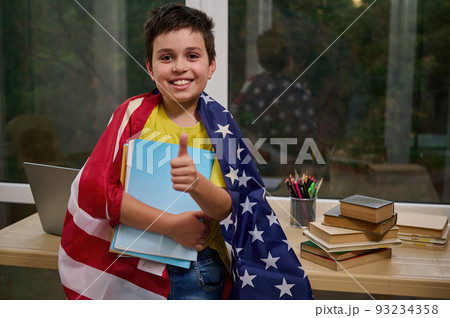 Happy Caucasian teenage boy, smart schoolchild, primary school student, wrapped in American flag, shows thumb up, holds textbooks, smiles looking at camera while standing against a desk with laptop Happy Caucasian teenage boy, smart schoolchild, primary school student, wrapped in American flag, shows thumb up, holds textbooks, smiles looking at camera while standing against a desk with laptop 93234358