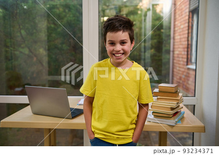 Portrait of a handsome Caucasian schoolboy, standing by a table with laptop, school supplies and stacked textbooks, cute smiling looking at camera. Back to school in the new semester of academic year Portrait of a handsome Caucasian schoolboy, standing by a table with laptop, school supplies and stacked textbooks, cute smiling looking at camera. Back to school in the new semester of academic year 93234371