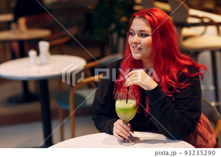 European plus size woman drink green smoothie in cafe. Young red pink haired body positive girl sitting at restaurant 93235290
