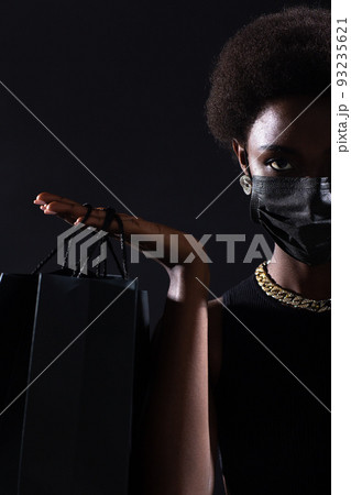 African american woman in black medical face mask holds black shopping bags. Safety on Black Friday concept. Covid-19 coronavirus protection. 93235621