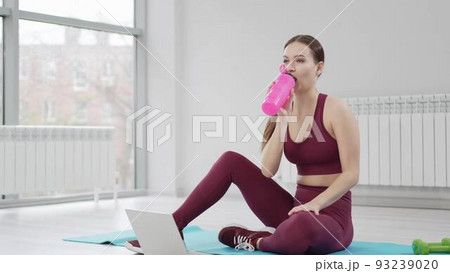 a young woman drinks water in a burgundy tracksuit with laptop on a mat in a gym a young woman drinks water in a burgundy tracksuit with laptop on a mat in a gym 93239020