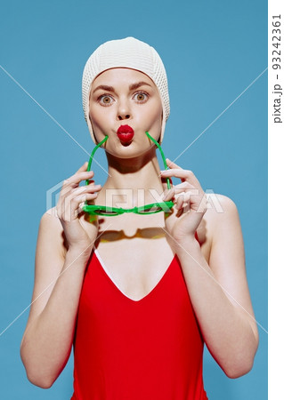 Playful woman in a swimming cap and a red swimsuit puts on green glasses folding her lips in a bow posing on a blue background 93242361