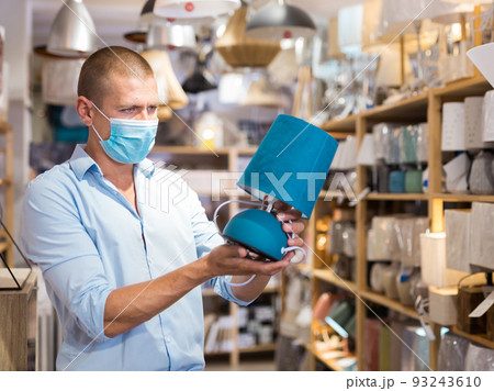 Man in protective mask is choosing new table lamp for her home in store 93243610