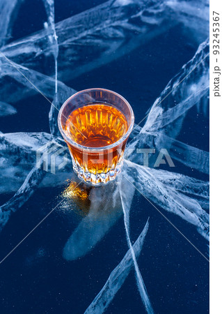 A glass of whiskey stands on beautiful ice with deep cracks. Orange liquid on a blue background. Top view from the side. Bizarre white deep cracks in the ice. Vertical. 93245367