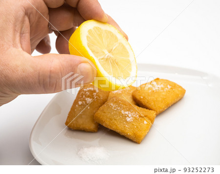 Hand squeezing a lemon on fresh panelle. Traditional Sicilian panelle on dish. Typical Sicilian street food. White Background. Hand squeezing a lemon on fresh panelle. Traditional Sicilian panelle on dish. Typical Sicilian street food. White Background. 93252273