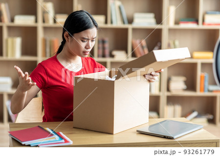 Frustrated disappointed young japanese lady opens cardboard box and sad for mistake with parcel 93261779