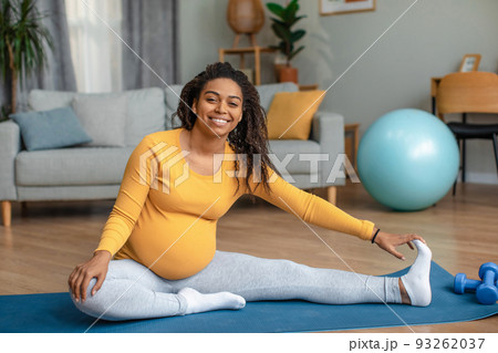 Happy young pregnant african american woman with big belly doing exercises and practicing yoga in living room Happy young pregnant african american woman with big belly doing exercises and practicing yoga in living room 93262037