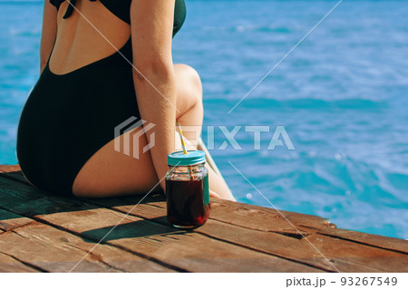 Attractive girl enjoys sea view on vacation. Woman in black swimsuit is sitting on edge of a wooden pier with a bottle of refreshing lemonade next to her. Tourism and travel. Copy space. Attractive girl enjoys sea view on vacation. Woman in black swimsuit is sitting on edge of a wooden pier with a bottle of refreshing lemonade next to her. Tourism and travel. Copy space. 93267549