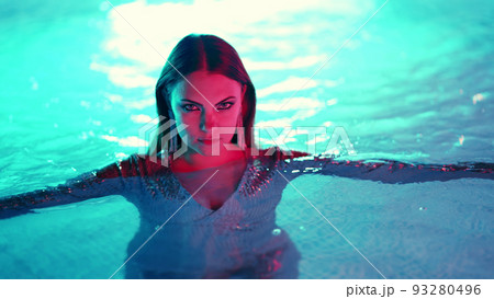 Beautiful woman posing in pool water under neon color light. Party, attractive chic in shining dress Beautiful woman posing in pool water under neon color light. Party, attractive chic in shining dress 93280496