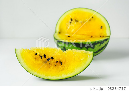 One slice of yellow watermelon in close-up, behind it a blurred oval of a striped giant berry in green rind, on a white background. Isolated slice of watermelon with yellow flesh on light surface. 93284757