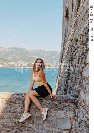 Beautiful girl sitting on a stone wall, in background is the blue sea, Budva, Montenegro. 93287039