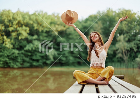 Hippie eco-activist woman traveler sits on a bridge by a lake with her arms outstretched with a hat and smiling sincerely Hippie eco-activist woman traveler sits on a bridge by a lake with her arms outstretched with a hat and smiling sincerely 93287498