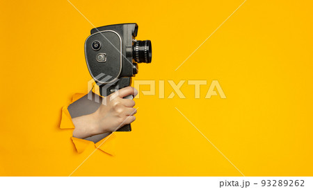 Hand holding small video old retro black camera 8 mm in paper torn hole in yellow background with copy space. Hand holding small video old retro black camera 8 mm in paper torn hole in yellow background with copy space. 93289262