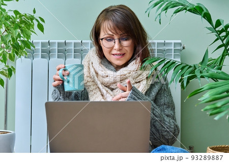 Middle aged female in warm clothes near heating radiator using laptop for video call Middle aged female in warm clothes near heating radiator using laptop for video call 93289887