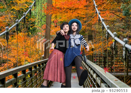 紅葉した自然の風景を背景に観光地で肩を組みながら、つり橋を渡る二人の女性旅行者 93290553