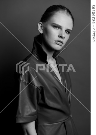 Seasonal Sale offer. Confidence blond young lady in trendy leather trench coat dress looks with blue eyes at camera presenting evening makeup posing isolated on olive green studio wall background Seasonal Sale offer. Confidence blond young lady in trendy leather trench coat dress looks with blue eyes at camera presenting evening makeup posing isolated on olive green studio wall background 93290595