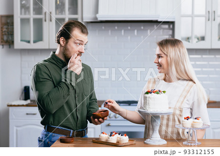 Pastry chef confectioner young caucasian woman with husband boyfriend man try taste of cake on kitchen table. Cakes cupcakes and sweet dessert 93312155