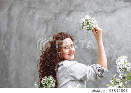 Girl with long curly brown hair in linen shirt holds blossoming branch of apple tree in hand against background of textured concrete wall. Spring flowering time. Copy SpaceIndependence Day of Ukraine 93312868
