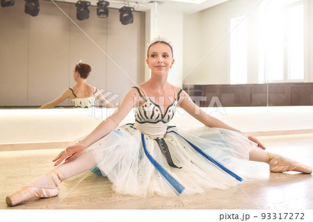 A young ballerina practicing ballet poses sitting against the mirror in the dance studio. A young ballerina practicing ballet poses sitting against the mirror in the dance studio. 93317272