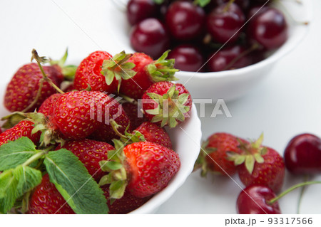 In the foreground is a white plate with strawberries and mint, behind it is a plate with cherries in close-up on a white background. High quality photo 93317566