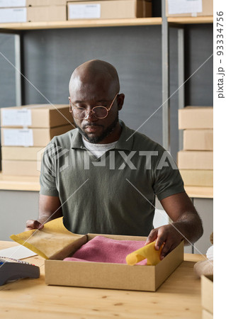 African warehouse worker packing order of customer in cardboard box while sitting at table in storage room African warehouse worker packing order of customer in cardboard box while sitting at table in storage room 93334756