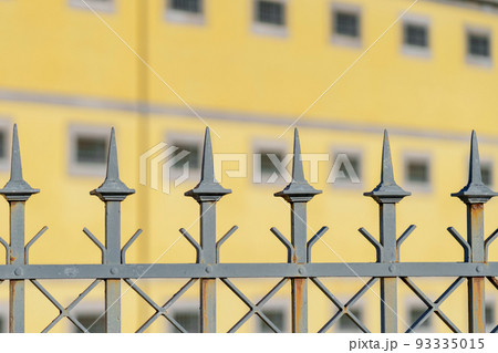 Metal fence with spikes of an old historical prison building with barred windows and yellow wall 93335015