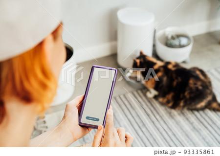 Female hands holding smartphone and using application to control cat food dispenser. Cat eating from smart feeder in cozy home interior on background. Home life with a pet. Healthy pet food diet. 93335861