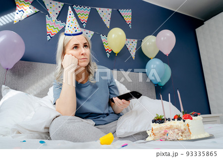 Sad woman in pajamas, party cap drinks champagne and watches TV, wrapped in a blanket while sitting on a bed in a decorated bedroom. Celebrates a birthday alone. Boring home party. Selective focus 93335865