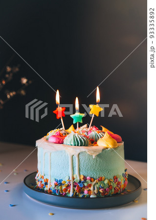 Colorful birthday cake with sprinkles and burning star shaped candles on a dark wall background. Festive birthday celebration, party. Vertical card. Selective focus, copy space. Colorful birthday cake with sprinkles and burning star shaped candles on a dark wall background. Festive birthday celebration, party. Vertical card. Selective focus, copy space. 93335892