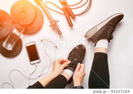 Young woman in sporting leggings laces sneakers, preparing for training. Accessories for sports on white background flat lay top view. Young woman in sporting leggings laces sneakers, preparing for training. Accessories for sports on white background flat lay top view. 93337104