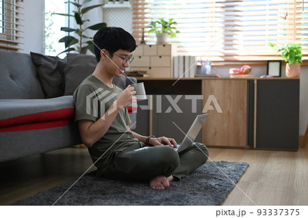Asian man in casual clothes sitting on carpet in cozy living room and browsing internet with laptop Asian man in casual clothes sitting on carpet in cozy living room and browsing internet with laptop 93337375