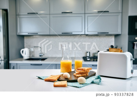 Toaster with fresh toast, eggs and glass of orange juice on a light kitchen table Toaster with fresh toast, eggs and glass of orange juice on a light kitchen table 93337828
