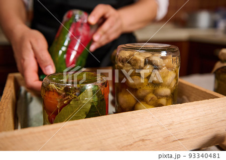 Selective focus on jars of canned vegetables stacked upside down in wooden box on blurred background of a housewife holding a can of chili peppers. Homemade preserved marinated fermented Vegetables 93340481
