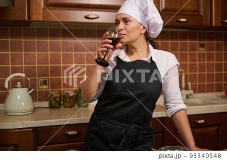 Beautiful dark-haired multi-ethnic confident woman, pleasant housewife wearing white chef's cap and black apron drinking red wine, enjoying cooking at home on her day-off. Domestic life, relaxation Beautiful dark-haired multi-ethnic confident woman, pleasant housewife wearing white chef's cap and black apron drinking red wine, enjoying cooking at home on her day-off. Domestic life, relaxation 93340548