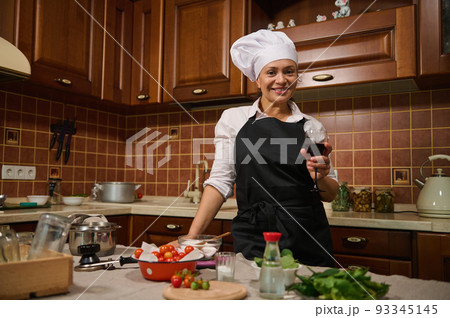Beautiful Hispanic housewife woman in white cap and back apron, holding a glass of red wine and smiling cutely at camera, standing in a cozy wooden home kitchen with ingredients on a kitchen island Beautiful Hispanic housewife woman in white cap and back apron, holding a glass of red wine and smiling cutely at camera, standing in a cozy wooden home kitchen with ingredients on a kitchen island 93345145