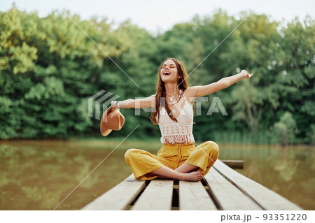 Hippie eco-activist woman traveler sits on a bridge by a lake with her arms outstretched with a hat and smiling sincerely Hippie eco-activist woman traveler sits on a bridge by a lake with her arms outstretched with a hat and smiling sincerely 93351220