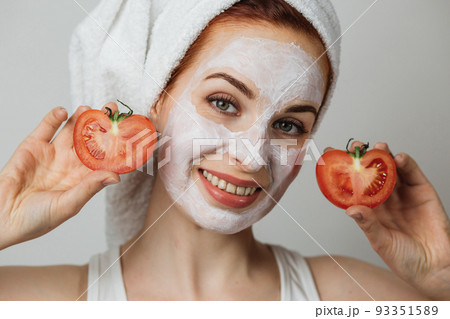 Caucasian young woman holding slices of tomato while posing in studio with white clay mask 93351589