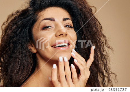 Happy tanned pretty curly Latin lady using facial cream after 30s to keep skin collagen elastin rate, hydrate and nourish it posing isolated on pastel beige background, smile at camera. Copy space 93351727